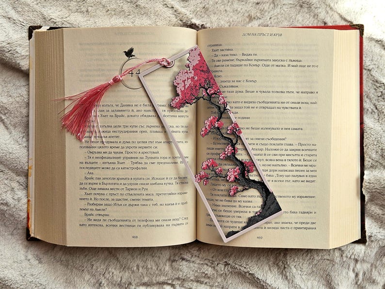 K&ouml;nnte beinhalten: Transparenter Acryl-Lesezeichen mit einem Kirschbl&uuml;ten-Design mit rosa Bl&uuml;ten und einem schwarzen Baumstamm. Das Lesezeichen hat eine rosa Quaste und liegt in einem offenen Buch.