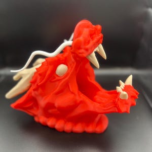 May include: A red 3D printed dragon head with white horns and a white tail. The dragon head is open-mouthed with visible teeth.