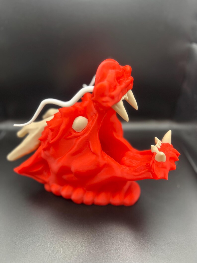 May include: A red 3D printed dragon head with white horns and a white tail. The dragon head is open-mouthed with visible teeth.