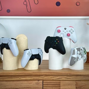 Pode incluir: Uma exibição de vários comandos de videojogos em branco, preto e com designs estampados, colocados em suportes decorativos de cor branco sujo. Os comandos estão dispostos numa superfície de madeira, mostrando acessórios de jogo.