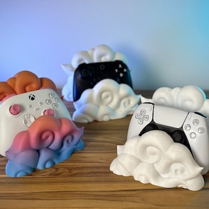 Pode incluir: Três suportes para controladores de jogos impressos em 3D de diferentes cores. Cada suporte tem a forma de uma nuvem e apresenta um esquema de cores diferente. Os suportes são projetados para segurar um controlador de jogos.