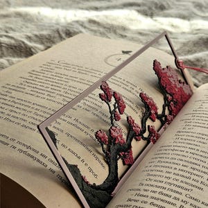 K&ouml;nnte beinhalten: Dekorativer Lesezeichen mit einem detaillierten, lasergeschnittenen Baumdesign mit roten Bl&uuml;ten und einem dunklen Stamm. Das Lesezeichen befindet sich in einem offenen Buch, mit einem roten Faden.