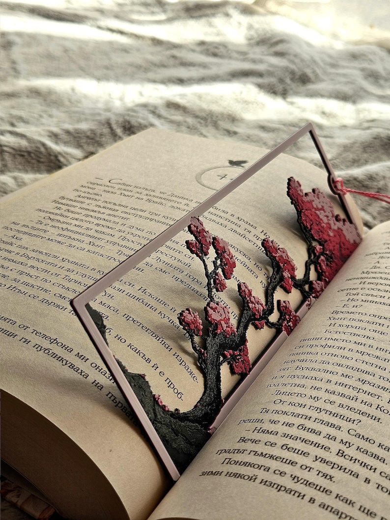 K&ouml;nnte beinhalten: Dekorativer Lesezeichen mit einem detaillierten, lasergeschnittenen Baumdesign mit roten Bl&uuml;ten und einem dunklen Stamm. Das Lesezeichen befindet sich in einem offenen Buch, mit einem roten Faden.