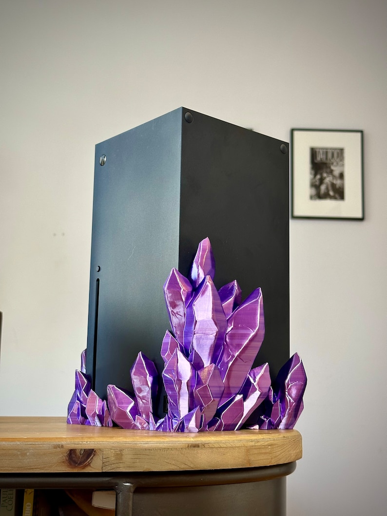 May include: A black gaming console sits on a purple crystal stand. The console is on a wooden table.