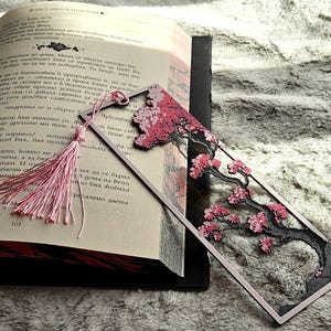 K&ouml;nnte beinhalten: Dekorativer Lesezeichen mit einem Kirschbl&uuml;ten-Design mit rosa Bl&uuml;ten und einem schwarzen Baumstamm. Das Lesezeichen hat eine rosa Quaste und liegt in einem offenen Buch.