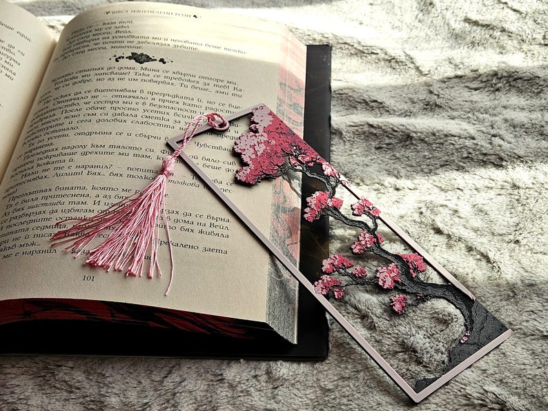 K&ouml;nnte beinhalten: Dekorativer Lesezeichen mit einem Kirschbl&uuml;ten-Design mit rosa Bl&uuml;ten und einem schwarzen Baumstamm. Das Lesezeichen hat eine rosa Quaste und liegt in einem offenen Buch.