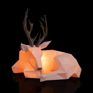 Pode incluir: Uma escultura geométrica de veado rosa claro com chifres castanhos escuros, repousando sobre uma superfície preta. O interior do veado brilha com uma luz laranja quente, criando um brilho ambiente suave. O design é moderno e minimalista.