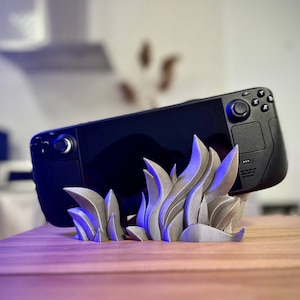 May include: A silver-coloured, 3D printed stand shaped like flames, holding a black handheld gaming device with a touch screen and buttons.
