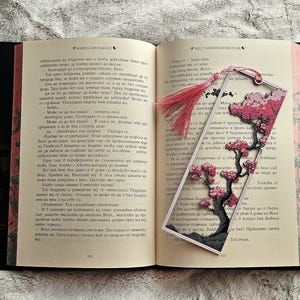 K&ouml;nnte beinhalten: Ein offenes Buch mit einem dekorativen Lesezeichen mit einem Kirschbl&uuml;ten-Design. Das Lesezeichen hat eine rosa Quaste und ein mehrschichtiges Papierdesign mit einem Baum und Blumen. Die Buchseiten sind sichtbar.
