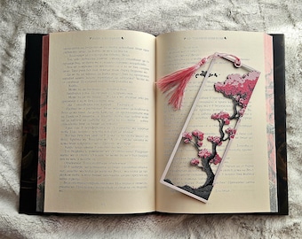 May include: An open book with a decorative bookmark featuring a cherry blossom design. The bookmark has a pink tassel and a layered paper design with a tree and flowers. The book pages are visible.