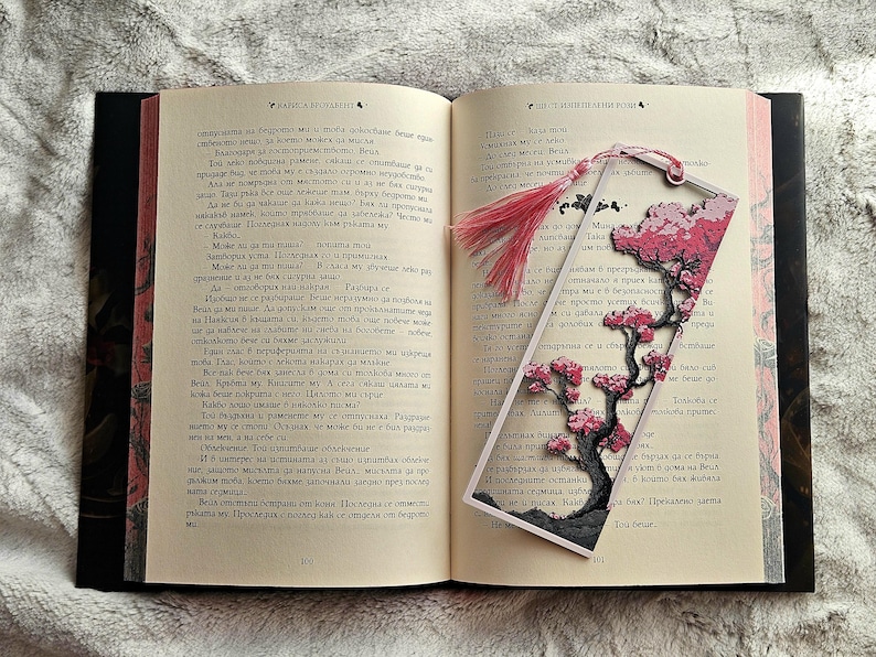 K&ouml;nnte beinhalten: Ein offenes Buch mit einem dekorativen Lesezeichen mit einem Kirschbl&uuml;ten-Design. Das Lesezeichen hat eine rosa Quaste und ein mehrschichtiges Papierdesign mit einem Baum und Blumen. Die Buchseiten sind sichtbar.