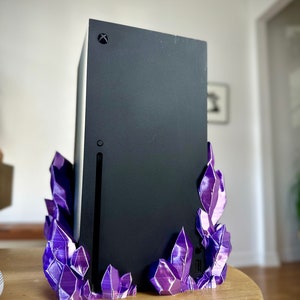 May include: A black Xbox Series X console on a purple crystal stand. The stand is made of 3D printed plastic and has a textured, crystalline appearance.