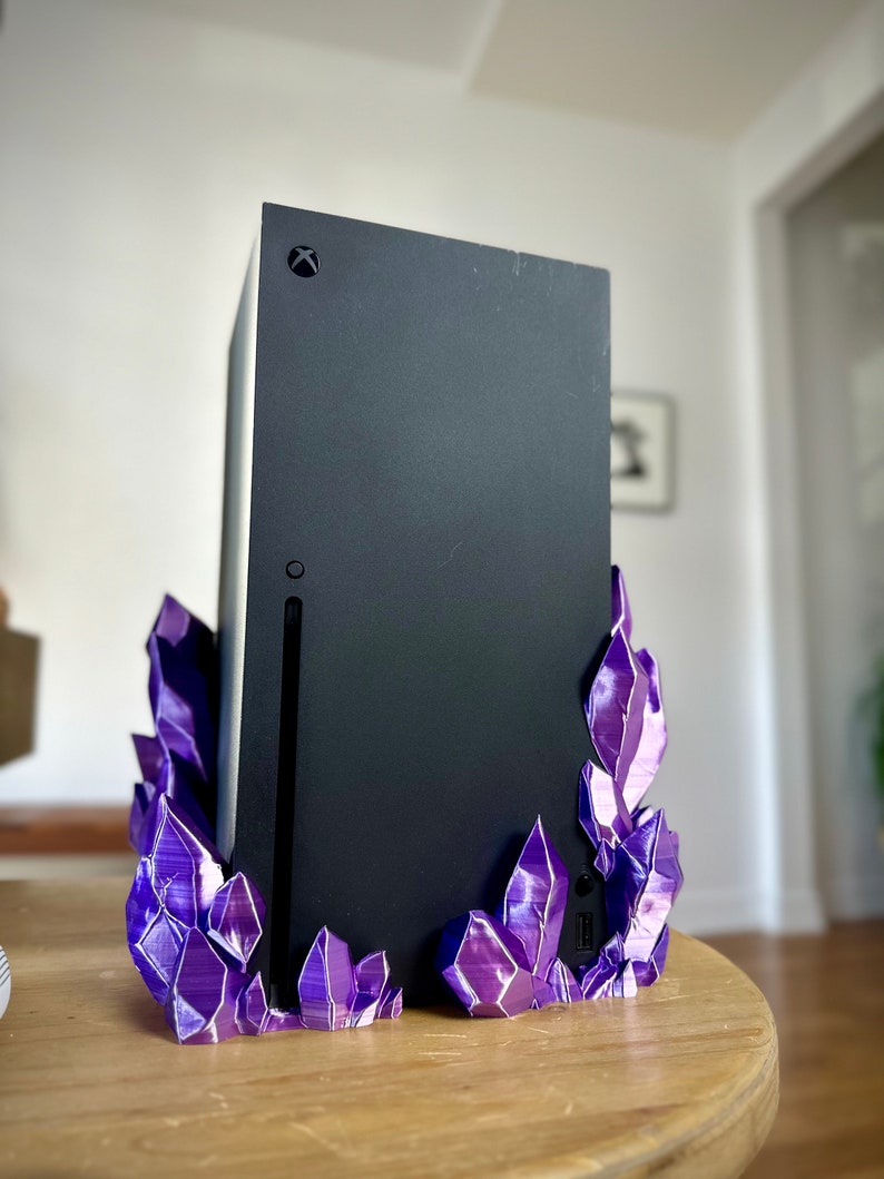 May include: A black Xbox Series X console on a purple crystal stand. The stand is made of 3D printed plastic and has a textured, crystalline appearance.