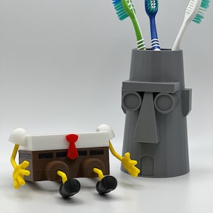 Pode incluir: Um suporte de escova de dentes cinzento em forma de cabeça com um nariz grande, contendo três escovas de dentes com cerdas verdes, azuis e brancas. Ao lado, um recipiente retangular castanho e branco com braços amarelos e sapatos pretos.