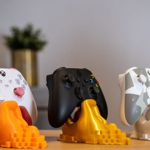 Könnte beinhalten: Drei Xbox-Controller in verschiedenen Farben werden auf Ständern präsentiert. Die Controller sind weiß, schwarz und grau mit einem weißen und grauen Muster. Die Ständer sind orange, gold und weiß.