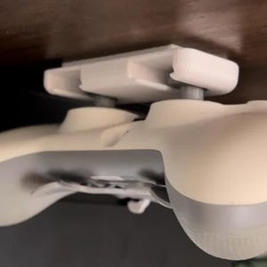 May include: A white video game controller is mounted to a white plastic bracket. The bracket is attached to a surface above the controller.