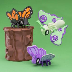May include: Three colorful insect figurines on a green background. One black insect with orange and black wings sits atop a brown tree stump. Two other insects have green, purple, and white details. The figurines are likely toys or decorative items.