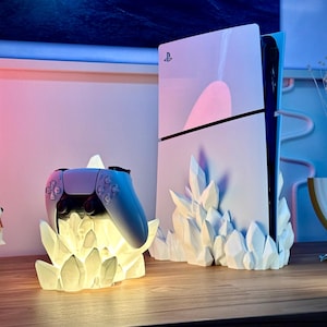 Może przedstawiać: Biała konsola PlayStation 5 z białym stojakiem w kształcie kryształu. Biały kontroler PlayStation 5 stoi na białym stojaku w kształcie kryształu.