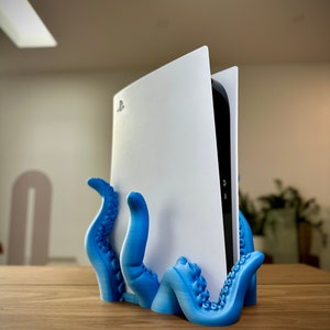 May include: A blue 3D printed octopus stand holding a white PlayStation 5 console. The console is standing upright with the PlayStation logo visible.