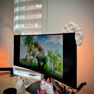 以下が含まれることがあります： 両側に白い雲の形をしたスタンドが付いた湾曲したコンピュータモニター。モニターには映画またはテレビ番組のシーンが表示されています。ビデオゲームコントローラーは、モニターの前にあるピンクとパープルのクリスタルベースの上に置かれています。