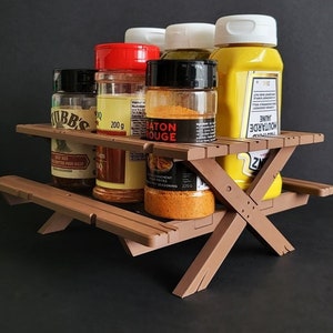 Pode incluir: Um suporte de especiarias de madeira em forma de mesa de piquenique com cinco frascos de especiarias de vidro. Os frascos estão cheios de várias especiarias, incluindo mostarda, temperos e ervas. Os frascos têm etiquetas com texto, incluindo "Baton Rouge" e "Moutarde Jaune".