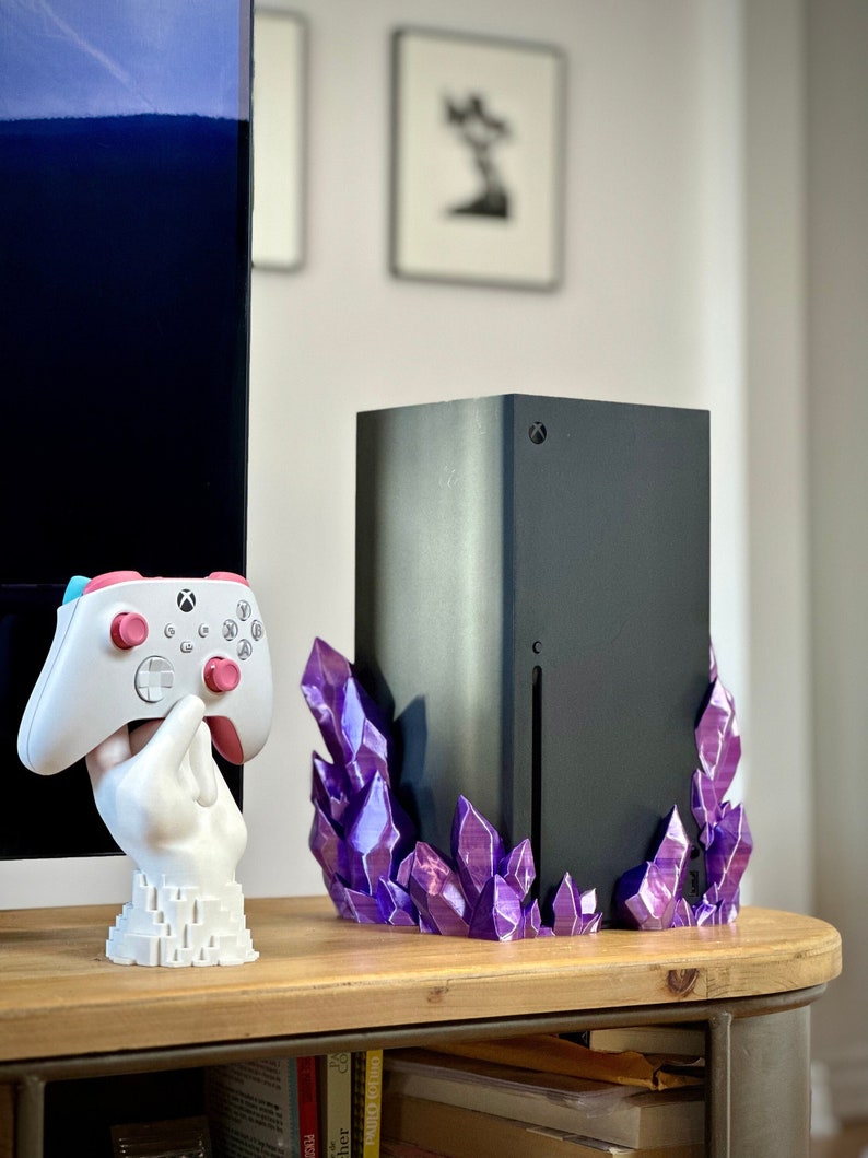 May include: A black gaming console with a purple crystal stand. The console is sitting on a wooden shelf with books underneath. A white hand holding a white and pink video game controller is to the left of the console.