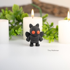 May include: A black Tiny Mothman figurine with orange eyes and wings stands between three lit white candles. Green foliage and a wooden branch are in the background. The text "Tiny Mothman" is at the bottom.