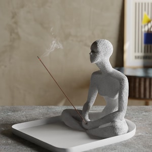 Op de afbeelding: Witte keramische wierookbrander in de vorm van een persoon die in meditatieve houding zit. De wierookstok brandt en er komt rook uit. De brander staat op een wit vierkant dienblad.
