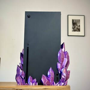 May include: A black gaming console with a purple crystal stand. The console has a black Xbox logo on the front. The stand is made of 3D printed plastic and has a shiny, iridescent finish.