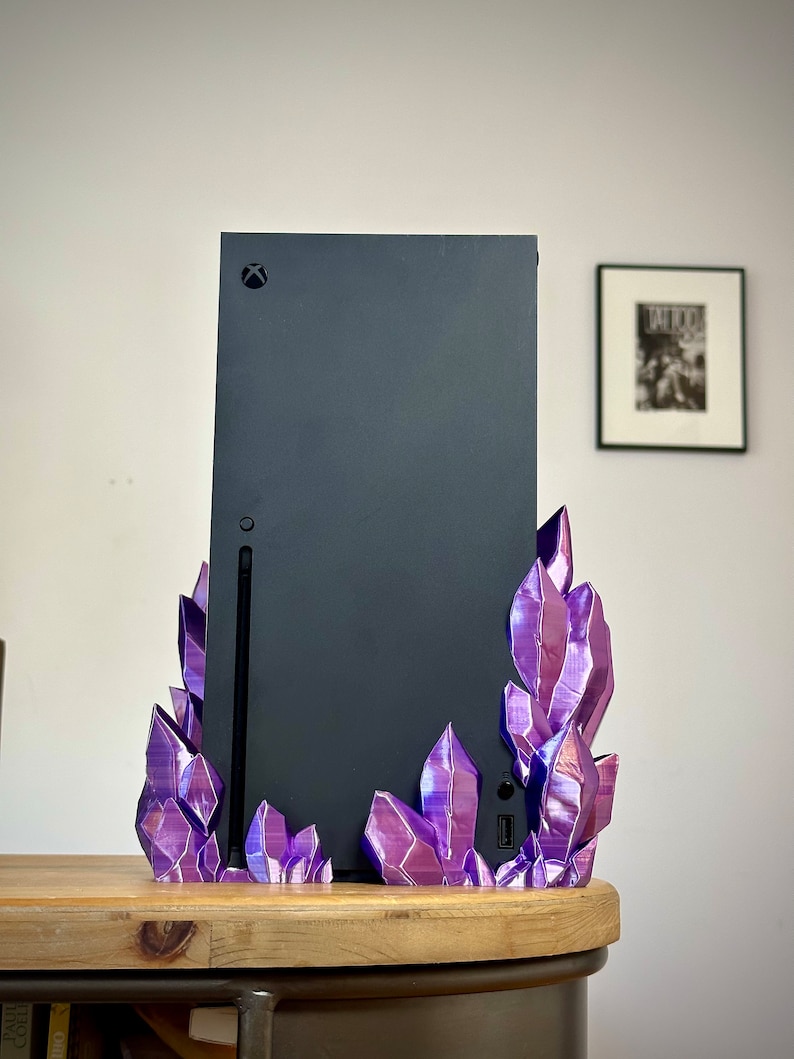 May include: A black gaming console with a purple crystal stand. The console has a black Xbox logo on the front. The stand is made of 3D printed plastic and has a shiny, iridescent finish.
