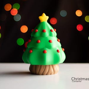 Könnte beinhalten: Eine grüne Weihnachtsbaum-Figur mit roten Kugeln und einem gelben Stern oben. Der Baum steht auf einer weißen Oberfläche mit einem verschwommenen Hintergrund aus bunten Lichtern.