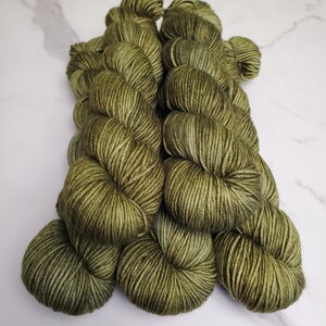 May include: A collection of olive green yarn skeins. The yarn has subtle variations in color, with hints of lighter and darker shades throughout. The skeins are neatly wound and arranged, suggesting a craft or textile project. The background is a white marble surface.