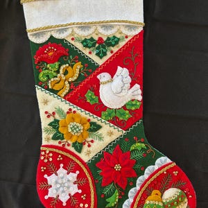 Könnte beinhalten: Handgefertigter Weihnachtsstrumpf in Rot, Grün und Weiß. Er zeigt gestickte Blumen- und Feiertagsmotive, darunter eine Taube, Poinsettien und einen Schneeflocke. Goldene Verzierungen und Akzente sorgen für einen festlichen Touch.