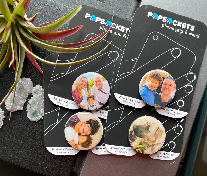 Puede incluir: Cuatro popsockets redondos para tel&eacute;fono con agarre y soporte, con diferentes im&aacute;genes. Las im&aacute;genes incluyen un retrato familiar, un padre y un hijo, una madre y un hijo, y un perro. Los popsockets son blancos con un contorno negro y el texto "iPhone X/8 users look inside" impreso en la parte posterior.