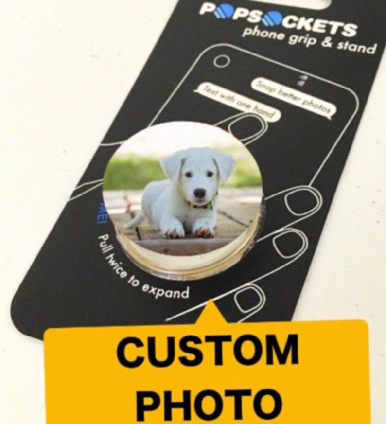 Puede incluir: Un cachorro blanco con un collar negro est&aacute; fotografiado sobre una superficie de madera. La imagen est&aacute; en una agarradera de tel&eacute;fono redonda y blanca con un borde negro. La agarradera de tel&eacute;fono est&aacute; etiquetada como "POPSOCKETS phone grip & stand" y "CUSTOM PHOTO".