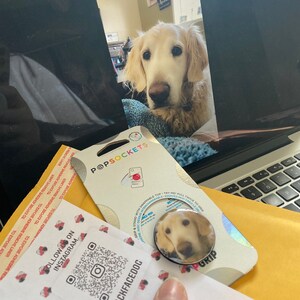 Puede incluir: Una agarradera de tel&eacute;fono blanca y dorada con una imagen de un perro golden retriever. La agarradera es redonda y tiene un anillo negro alrededor del borde. El texto "BLACKFACEDOG" est&aacute; impreso en la tarjeta blanca debajo de la agarradera. El texto "FOLLOW US ON INSTAGRAM" est&aacute; impreso en la tarjeta blanca encima de la agarradera. Un c&oacute;digo QR est&aacute; impreso en la tarjeta blanca.