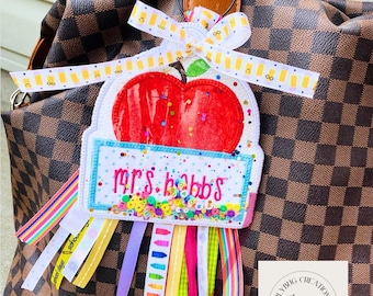 May include: A personalized teacher bag tag featuring a red apple design with the name "Mrs. Hobbs" on a blue and white label. The tag includes colorful ribbons and a bow, attached to a brown and tan checkered handbag.
