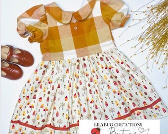 May include: A children's dress with a plaid bodice in shades of yellow, white, and tan, and a skirt featuring a repeating pattern of houses and trees on a cream background. The dress has a Peter Pan collar and short sleeves. Brown Mary Jane shoes are visible.