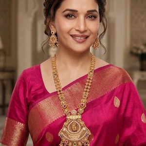 May include: A long gold necklace featuring an intricate circular pendant with a central deity figure, accented by small red and green stones and dangling gold beads. Matching gold drop earrings with similar stone details and filigree work.