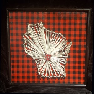 May include: A framed string art piece depicting the state of Wisconsin. The artwork features a white string outline of the state over a red and black buffalo plaid background. A red heart is in the center of the state.