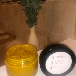 May include: A jar of turmeric honey body scrub with a black lid. The jar is filled with a bright yellow, thick scrub. The lid has a white label with the text "Turmeric Honey" and "Body Polish & Body Mask".