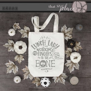 K&ouml;nnte beinhalten: Eine wei&szlig;e Tragetasche mit einem grauen Design, auf dem "Lunch Lady Working My Fingers To The Bone" steht, mit einer Skeletthand, die ein Shaka-Zeichen macht. Die Tasche ist von Herbstbl&auml;ttern und wei&szlig;en K&uuml;rbissen umgeben.