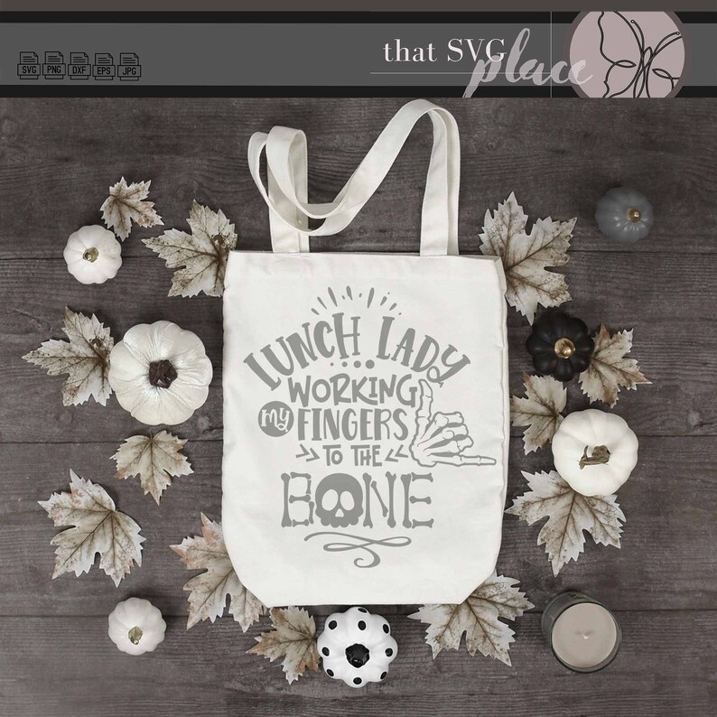 K&ouml;nnte beinhalten: Eine wei&szlig;e Tragetasche mit einem grauen Design, auf dem "Lunch Lady Working My Fingers To The Bone" steht, mit einer Skeletthand, die ein Shaka-Zeichen macht. Die Tasche ist von Herbstbl&auml;ttern und wei&szlig;en K&uuml;rbissen umgeben.