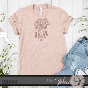 Op de afbeelding: Een lichtroze T-shirt met een roosgouden wolfskop in een droomvangerontwerp. Het ontwerp is gecentreerd op het shirt.