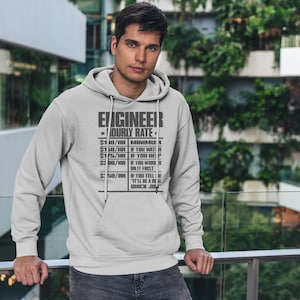 Op de afbeelding: Een grijs hoodie met de tekst "ENGINEER *HOURLY RATE*" en een lijst met uurtarieven voor verschillende taken. De tekst is zwart en de achtergrond is grijs.