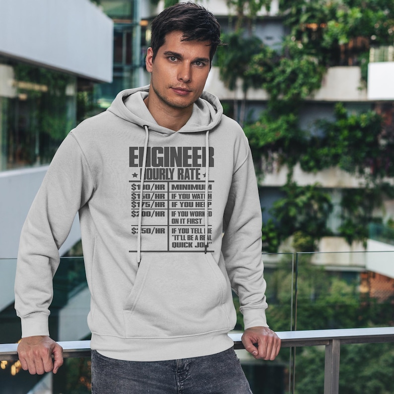 Op de afbeelding: Een grijs hoodie met de tekst "ENGINEER *HOURLY RATE*" en een lijst met uurtarieven voor verschillende taken. De tekst is zwart en de achtergrond is grijs.
