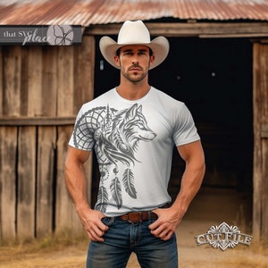 Op de afbeelding: Een wit T-shirt met een grijs wolfontwerp en een droomvanger met veren. De wolf kijkt naar rechts en de droomvanger bevindt zich aan de linkerkant van het shirt. Het shirt wordt gedragen door iemand die een witte cowboyhoed en een blauwe jeans draagt.