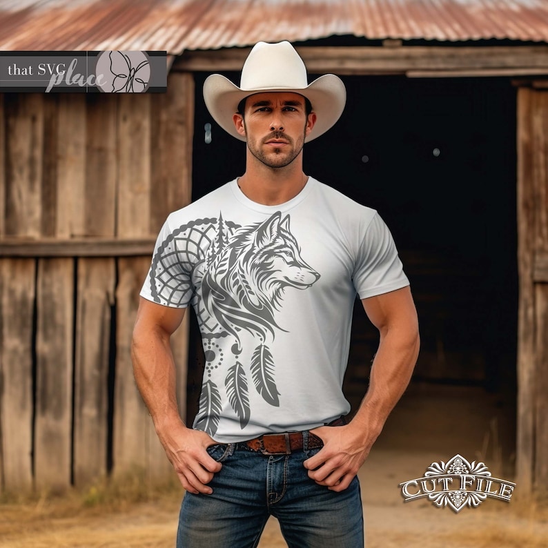 Op de afbeelding: Een wit T-shirt met een grijs wolfontwerp en een droomvanger met veren. De wolf kijkt naar rechts en de droomvanger bevindt zich aan de linkerkant van het shirt. Het shirt wordt gedragen door iemand die een witte cowboyhoed en een blauwe jeans draagt.