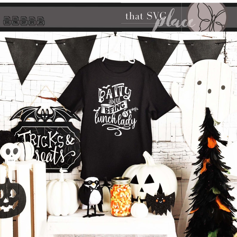 K&ouml;nnte beinhalten: Schwarzes T-Shirt mit wei&szlig;em Text, der "Batty about being a lunch lady" lautet. Das Shirt wird auf einem Regal mit Halloween-Dekorationen pr&auml;sentiert, darunter ein schwarz-wei&szlig;es Schild mit der Aufschrift "Tricks & Treats", eine schwarz-wei&szlig;e Fledermaus, ein wei&szlig;er K&uuml;rbis, ein schwarz-wei&szlig;er Pinguin und ein Glas mit Maisbonbons.