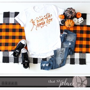 K&ouml;nnte beinhalten: Wei&szlig;es T-Shirt mit einem Skelett-Motiv und dem Text "Bone-afide Lunch Lady". Das Shirt ist auf einer karierten orange-schwarzen Decke mit einem Paar schwarzer Turnschuhe, einer blauen Jeans und kleinen orangefarbenen K&uuml;rbissen abgebildet.